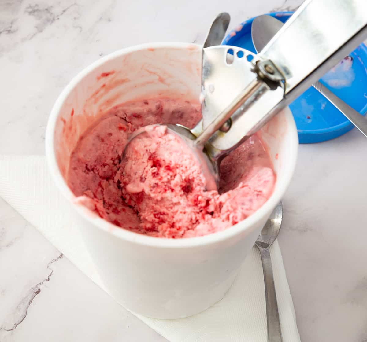 An ice cream tub with raspberry ice cream and scoop.