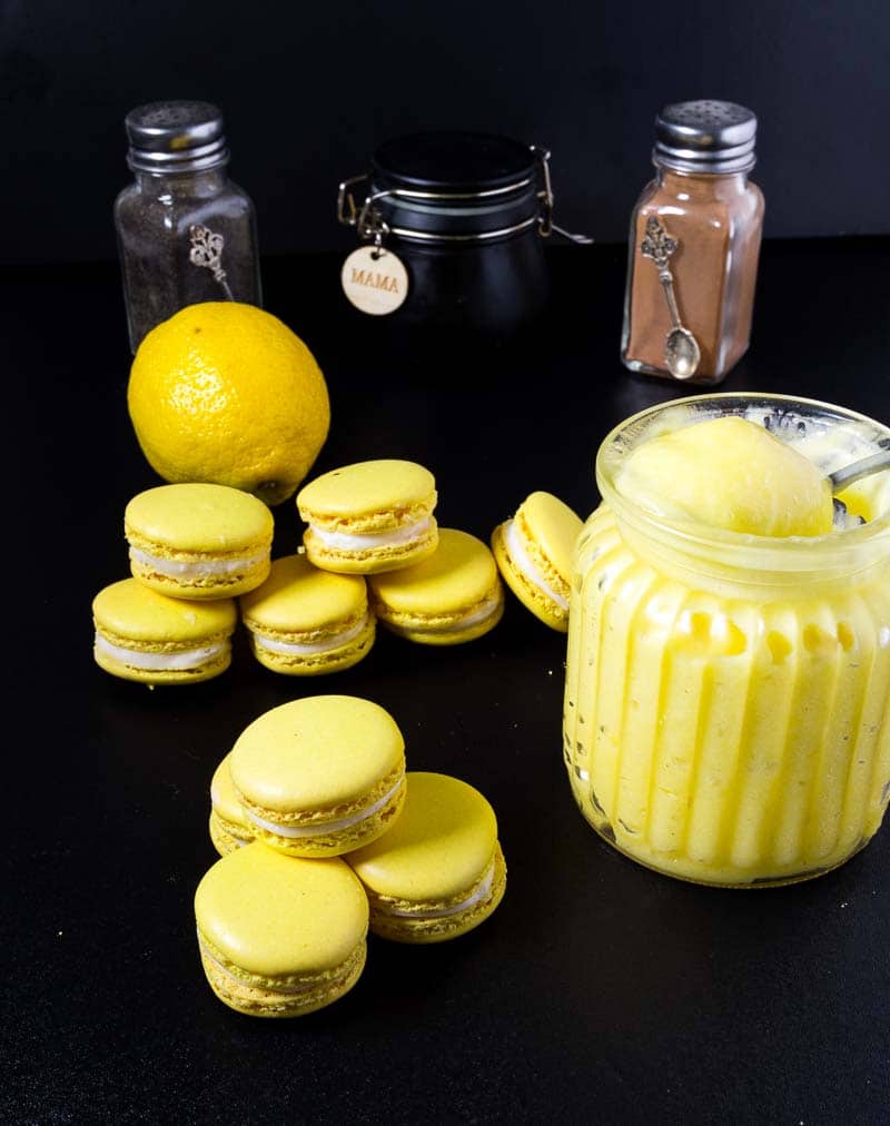 French Macarons are on-trend all year round. These lemon French macarons filled with lemon curd and Swiss meringue buttercream are especially refreshing and light. This simple, easy and effortless recipe uses my full proof macaron recipe as a base so you can make these successfully over and over again. People are going to think you are a macaron master baker!! A Pinterest Optimized image for Lemon French Macarons fill with homemade lemon curd and Swiss meringue buttercream using my full proof french macaron recipe