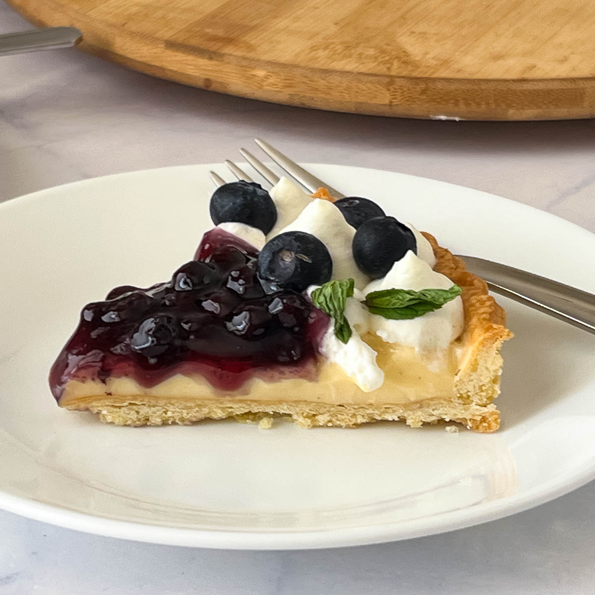 This blueberry tart is an absolute classic and luxurious dessert with rich, creamy vanilla pastry cream in a buttery short crust pastry, topped with a delicious blueberry compote and garnished with fresh blueberries. A simple, easy and effortless recipe that can be done a head of time. A slice of blueberry tart on the plate.