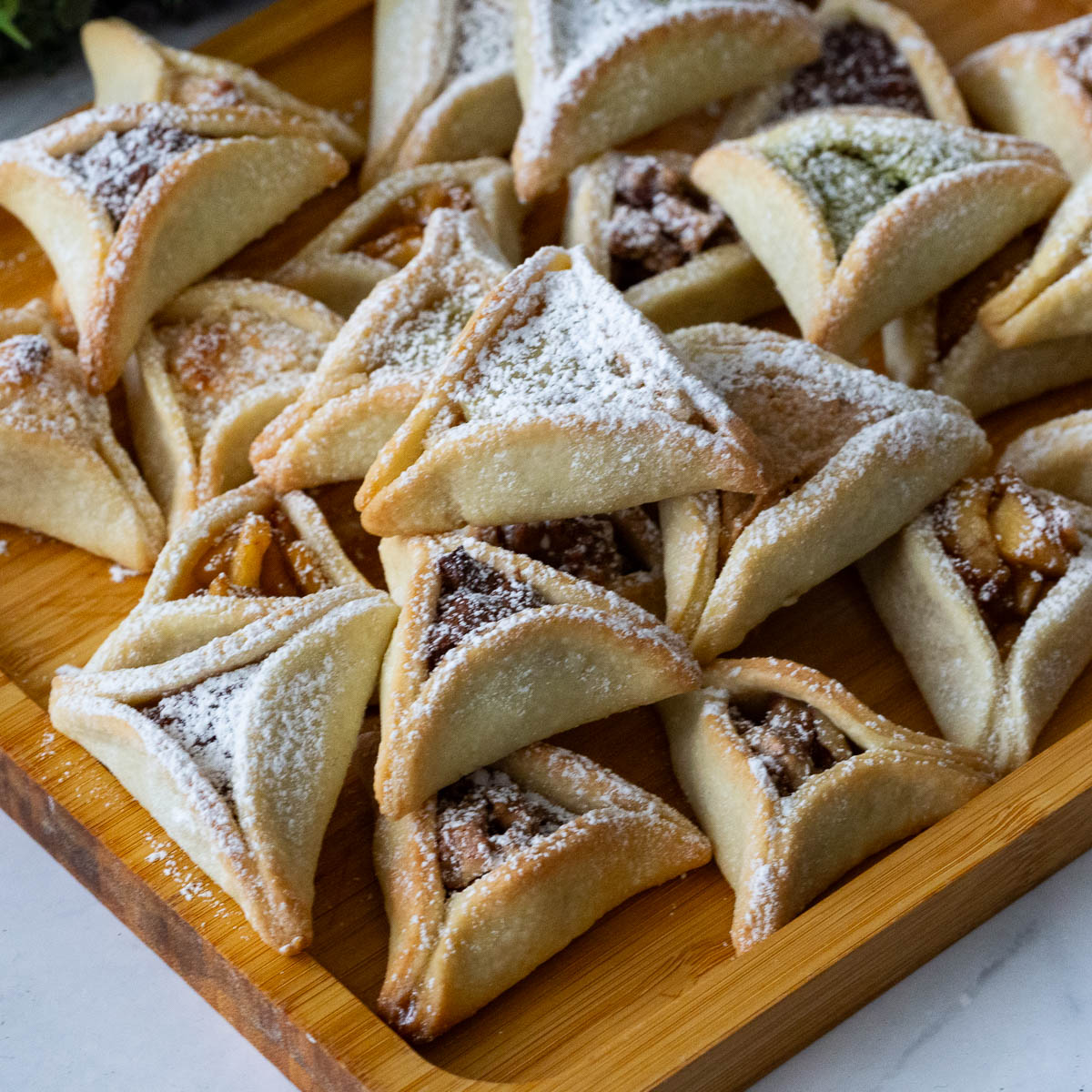 A Purim party is incomplete without Purim cookies and these dates Purim cookies are an absolute addiction. This simple, easy and effortless recipe for festive purim cookies also called hamentaschen or oznei haman are filled with naturally sweet dates and pecans then folded in a short bread cookie dough to get a traditional triangle shape. A stack of purim cookies on the table,