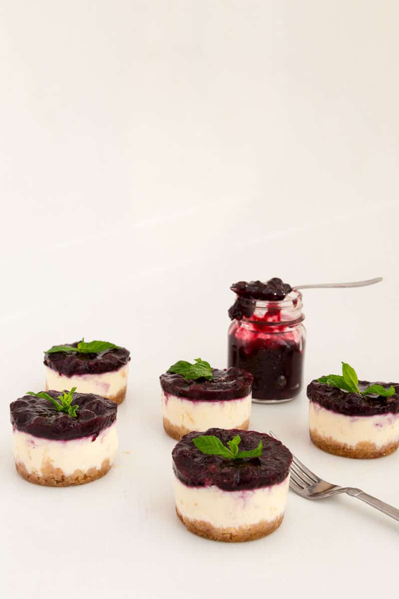Nothing beats cheesecake for dessert any time of the year. This simple, easy and effortless recipe for mini blueberry cheesecakes is perfect for entertaining or when you need the mini versions of the classic. A custard-based cheese batter baked to perfection then topped with luxurious blueberry filling is a perfect mini treat at the end of any meal. A Pinterest optimized image for Mini Blueberry Cheesecakes recipe showing few mini cheesecakes with a jar of fresh blueberry filing