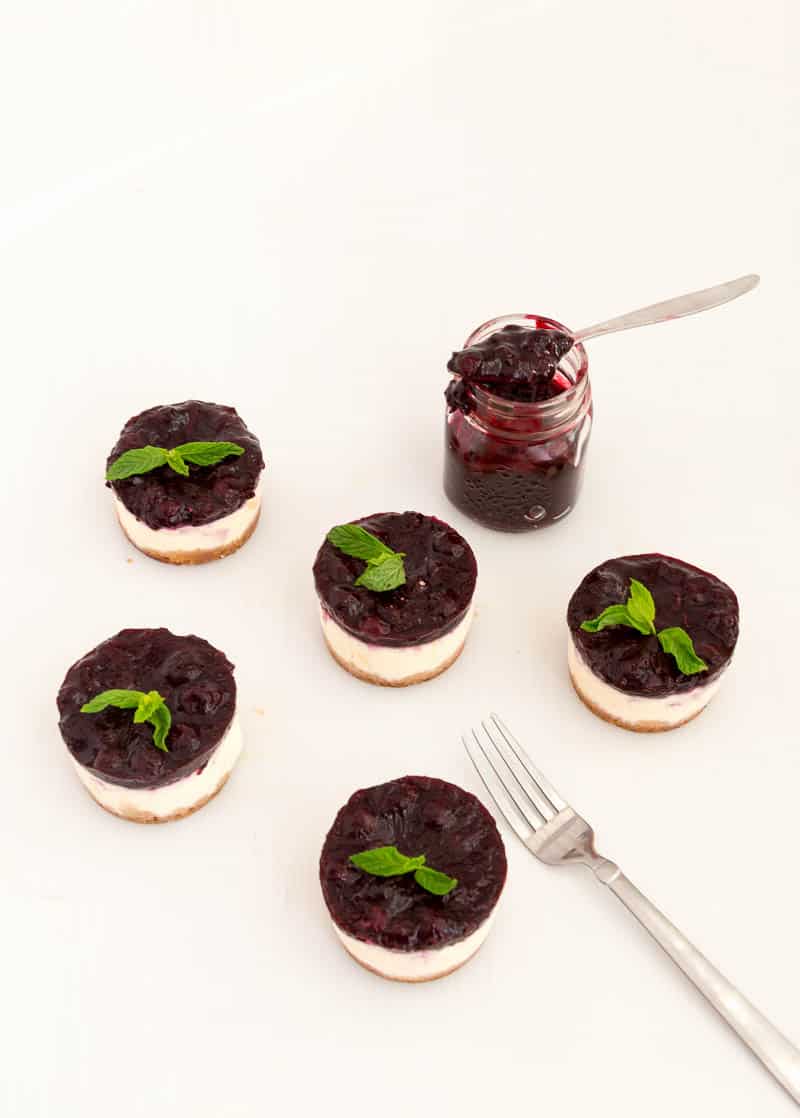 Nothing beats cheesecake for dessert any time of the year. This simple, easy and effortless recipe for mini blueberry cheesecakes is perfect for entertaining or when you need the mini versions of the classic. A custard-based cheese batter baked to perfection then topped with luxurious blueberry filling is a perfect mini treat at the end of any meal. A Pinterest optimized image for Mini Blueberry Cheesecakes recipe showing few mini cheesecakes with a jar of fresh blueberry filing