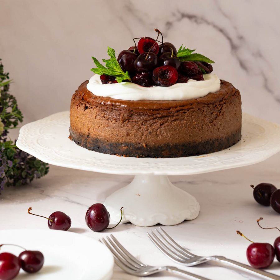 A baked chocolate cheesecake with cherry topping on a cake stand.