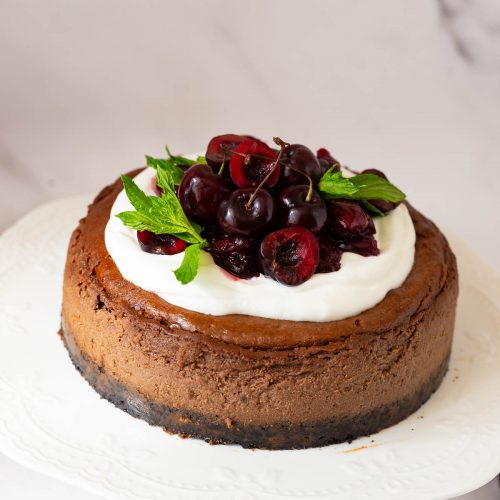 A chocolate cheesecake with whipped cream and cherry filling on a cake stand.