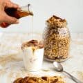 A person pouring maple syrup to glass full of yogurt and granola.