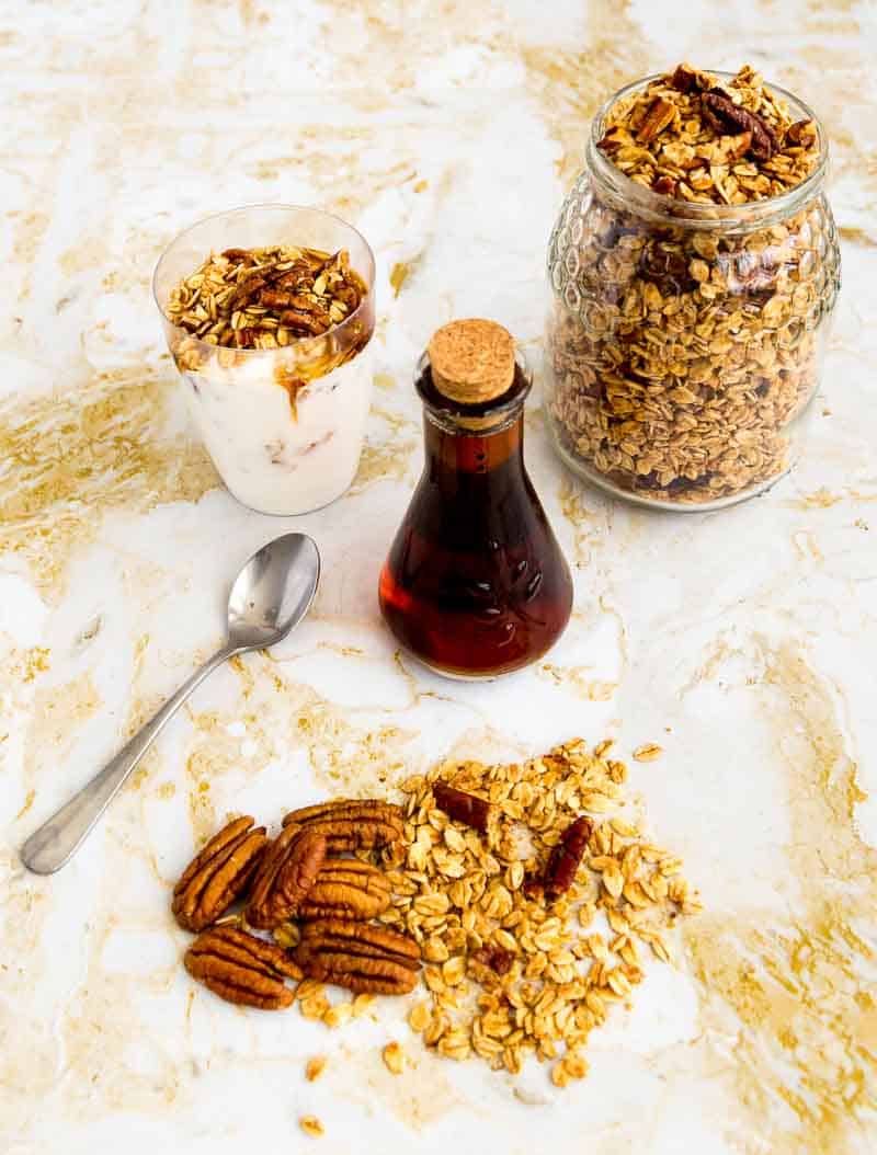 A glass full of yogurt and granola next to a jar full of granola and a bottle of maple syrup.