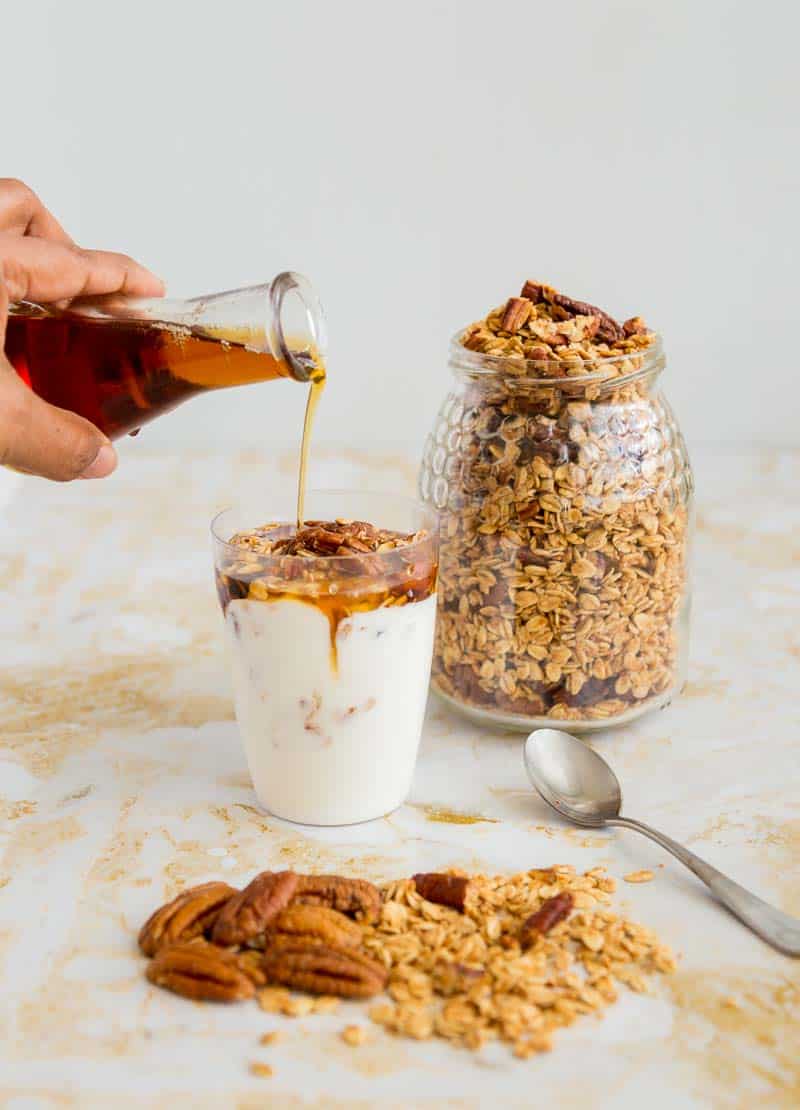 A person pouring maple syrup into glass full of yogurt and granola next to a jar full of granola.