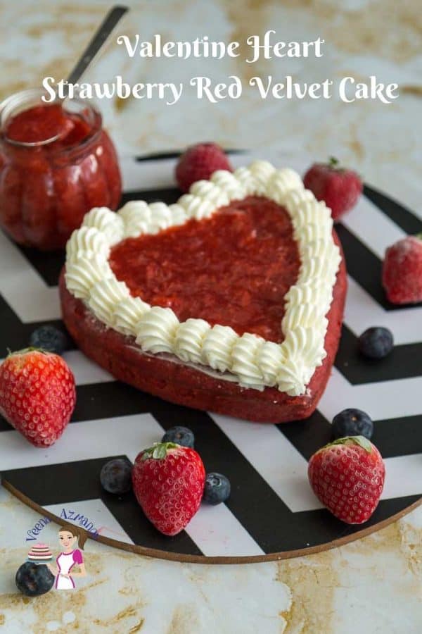 A red heart-shaped cake on a round board next to fresh strawberries.