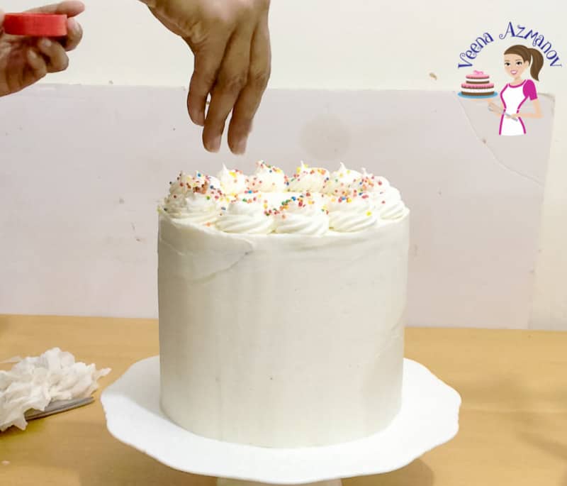 A person decorating a birthday cake with white frosting on a cake stand.