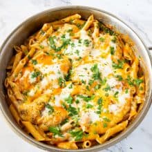 A skillet with pasta topped with chicken breast and cheese.
