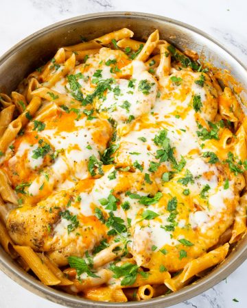 A skillet with pasta topped with chicken breast and cheese.
