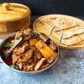 Lamb curry in a metal bowl next to chapatis on a wooden tray.