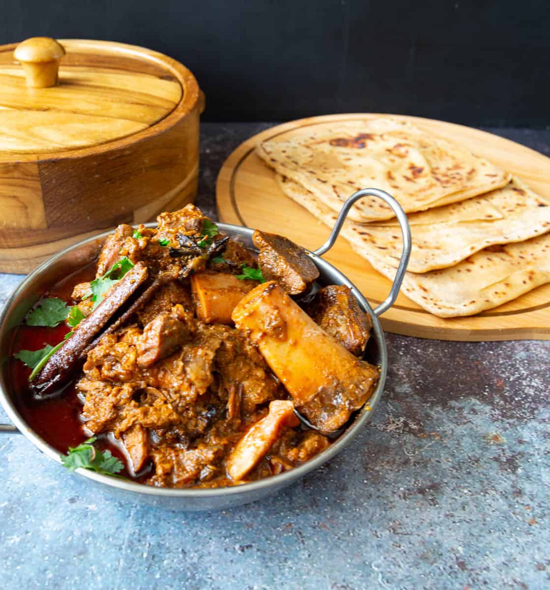 Lamb curry in a metal bowl next to chapatis on a wooden tray.