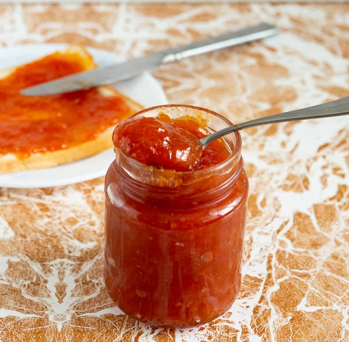 A jar of apricot peach jam and a slice of bread with jam on a plate with a knife.