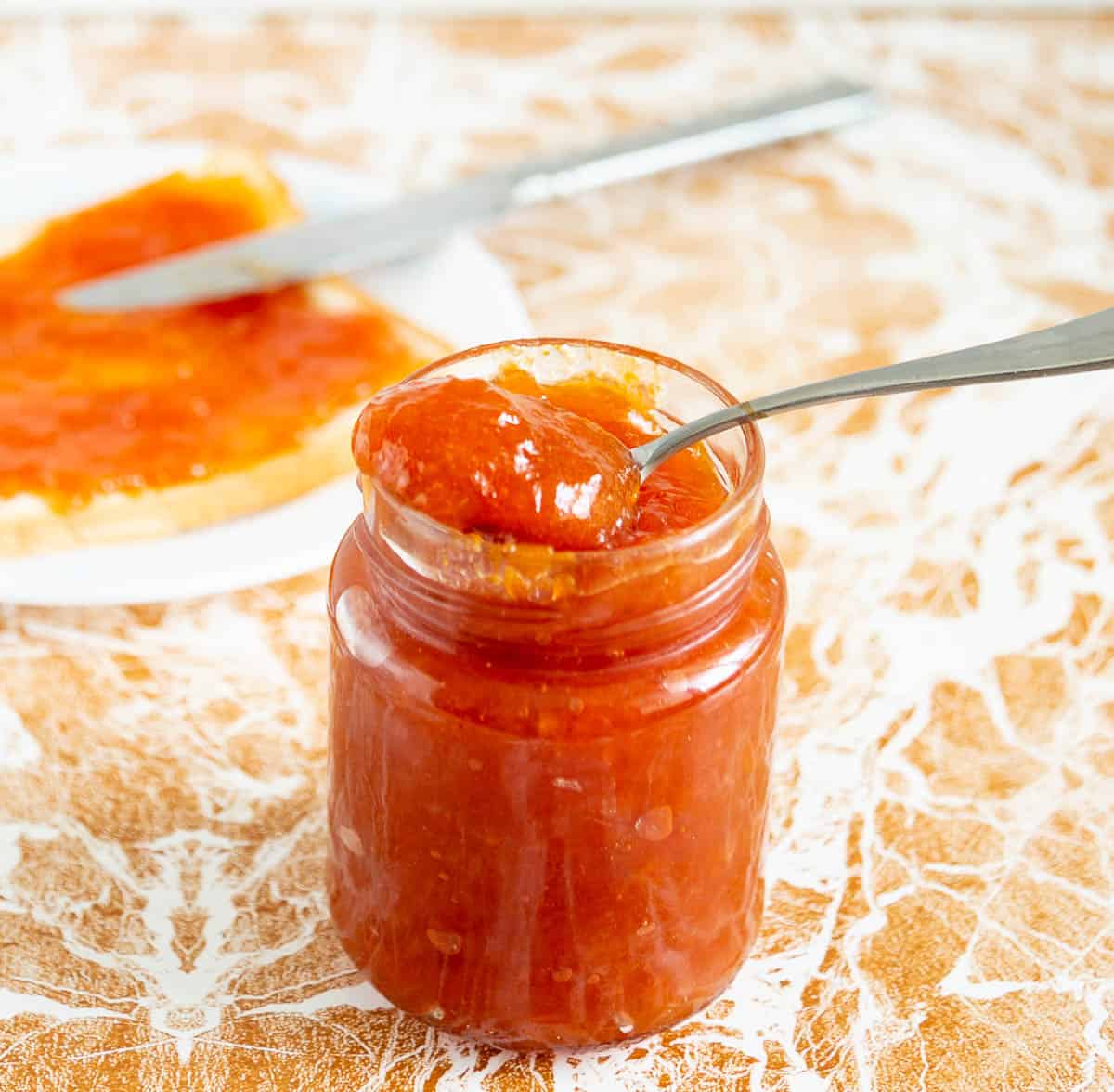 A jar of apricot peach jam and a slice of bread with jam on a plate with a knife.