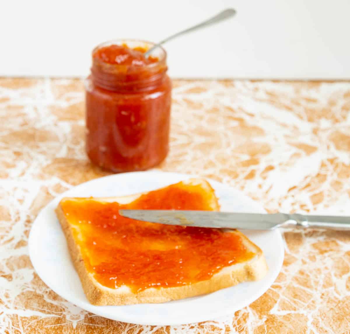 A slice of bread with apricot peach jam on a plate with a knife, and a jar of jam in the background.