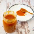 A glass jar filled with tomato sauce next to a plate with a spoon full of tomato sauce.