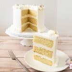 A slice of vanilla layer cake on a plate, with the rest of the cake on a cake stand.