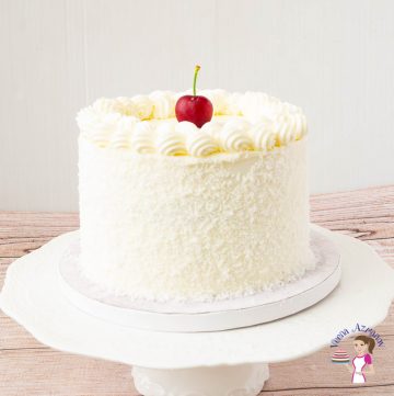 A coconut cake on a cake stand with a cherry on top.