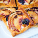 Blueberry peach puff pastry tart with cream cheese filling served on a white plate.