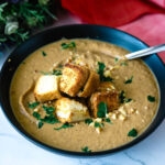 A black bowl with roasted cauliflower soup, croutons, and garnish.