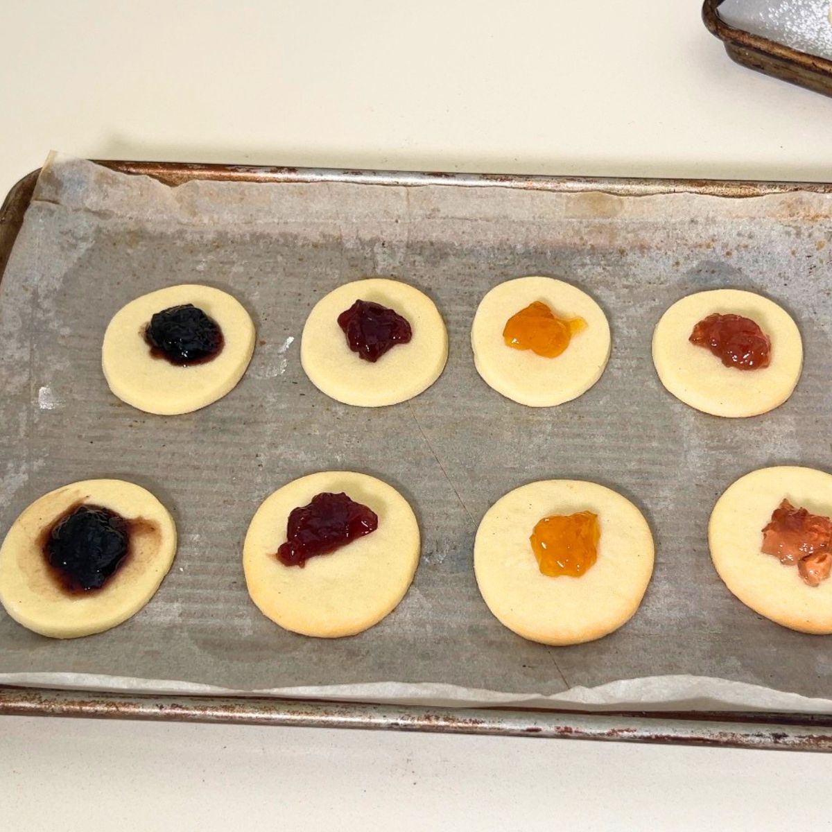 Bottom of sandwich cookies with jam on baking tray.